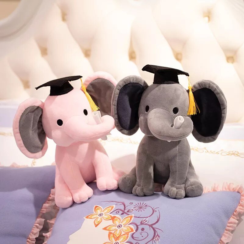 Two plush elephant toys, one pink and one gray, wearing graduation caps on a floral-patterned pillow.