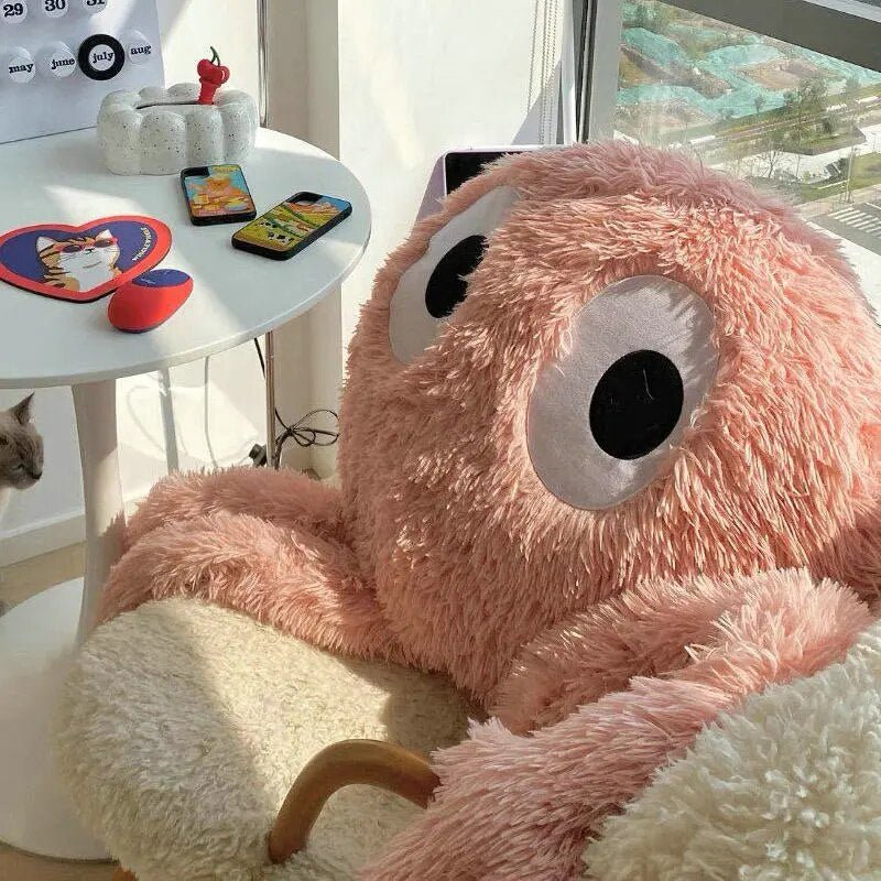 Large pink plush toy with googly eyes in a room with a desk and window.