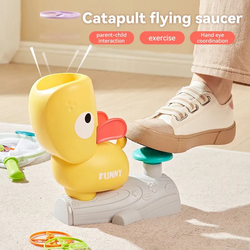 Yellow duck-shaped catapult toy with a foot on a platform, labeled 'Catapult flying saucer', on a light wooden floor.