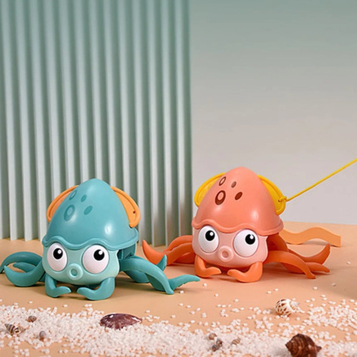 Two toy octopuses, one blue and one pink, on a sandy surface.