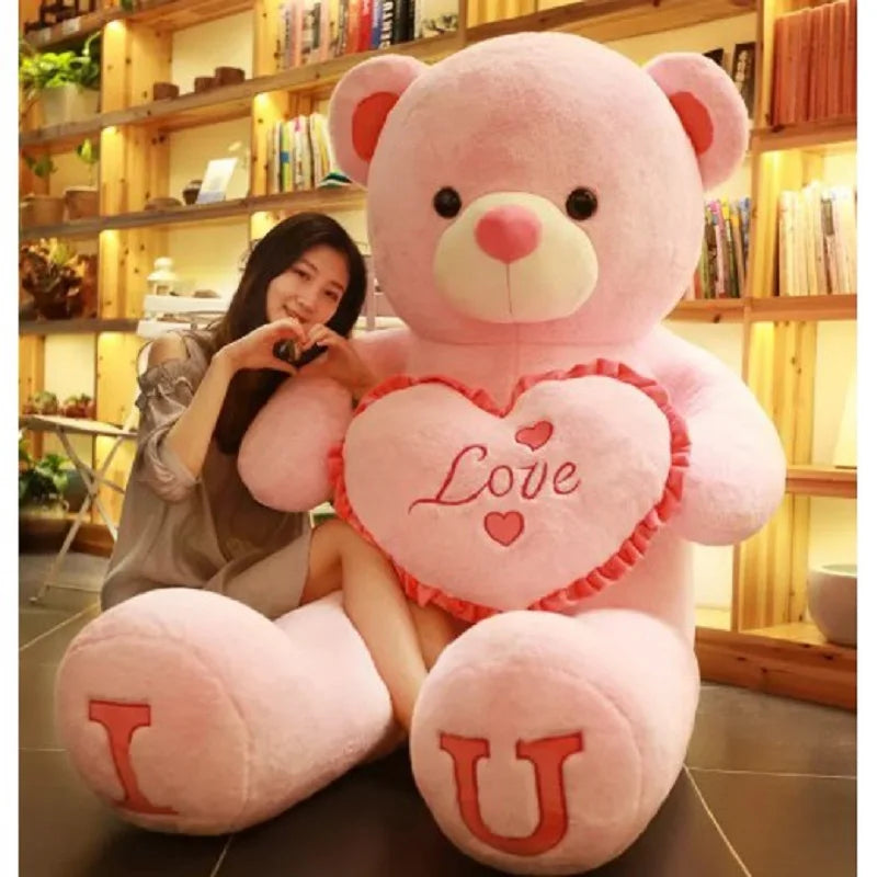 Person holding a large pink teddy bear with a heart that says 'I Love U' in a room with wooden shelves.