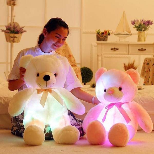 Two glowing teddy bears, one white and one pink, being held by a person in a bedroom setting.