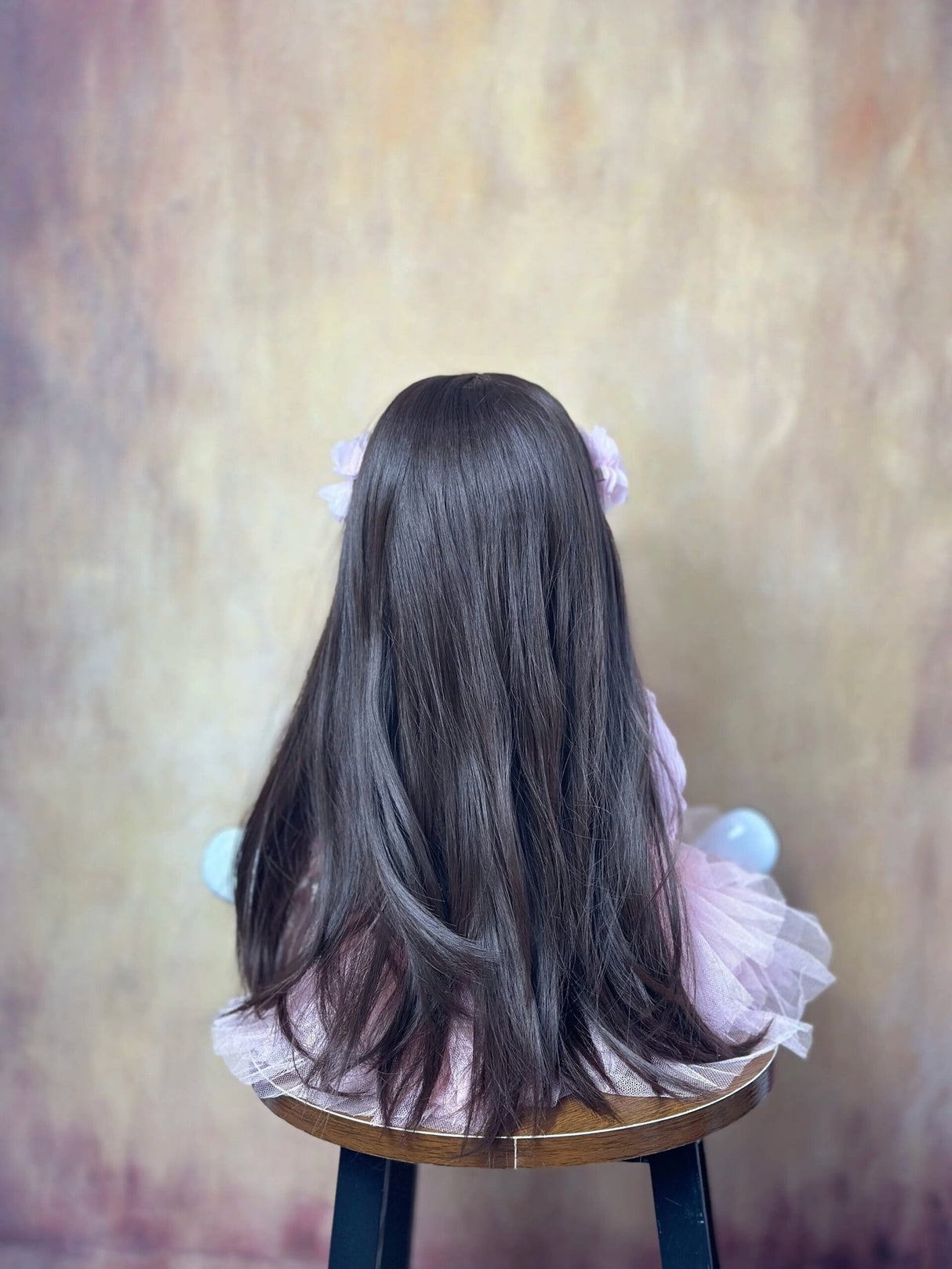 Back view of a doll with long dark hair sitting on a stool against a textured wall.