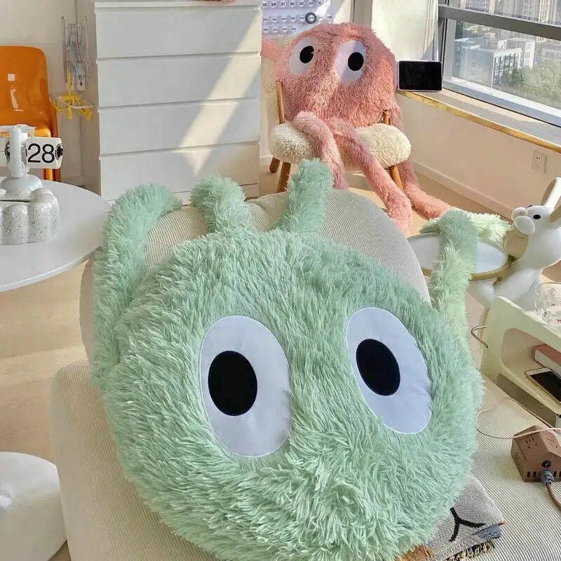 Green plush toy with googly eyes on a chair in a room with another plush toy.