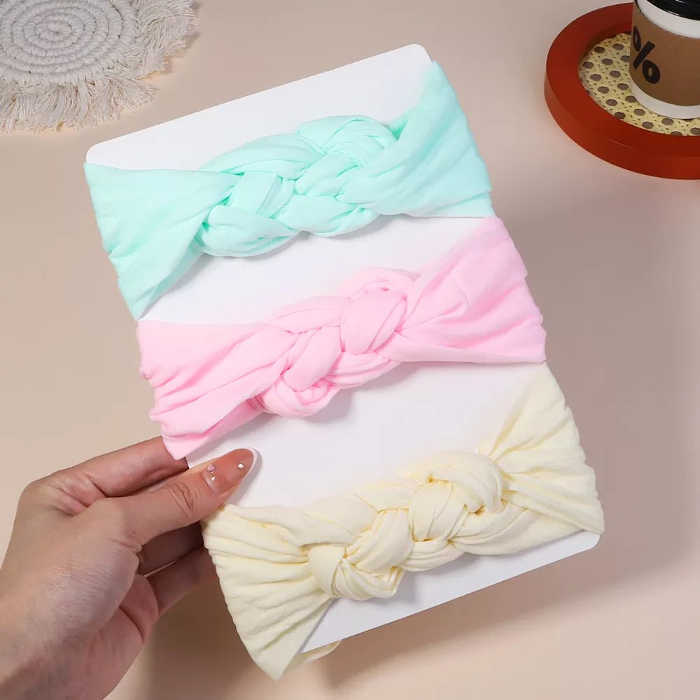 Three pastel-colored knotted headbands on a white surface with a hand holding the top one.