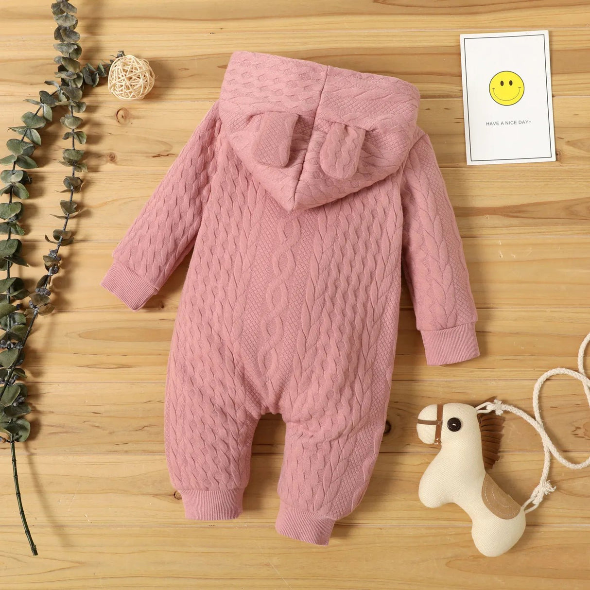Pink baby romper with cable knit pattern on a wooden surface with decorative elements.