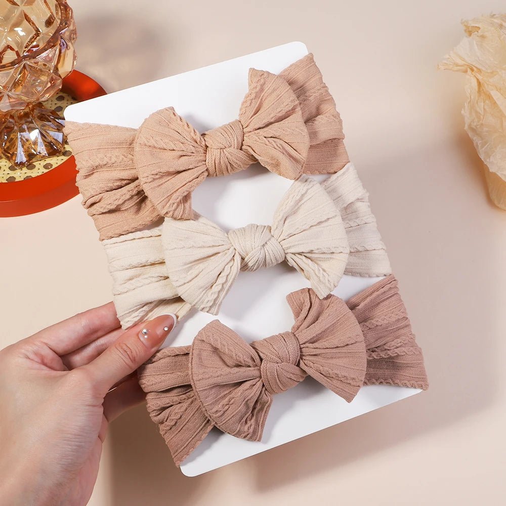 Three fabric hair bows in beige, pink, and brown on a white tray with a hand holding one of them.
