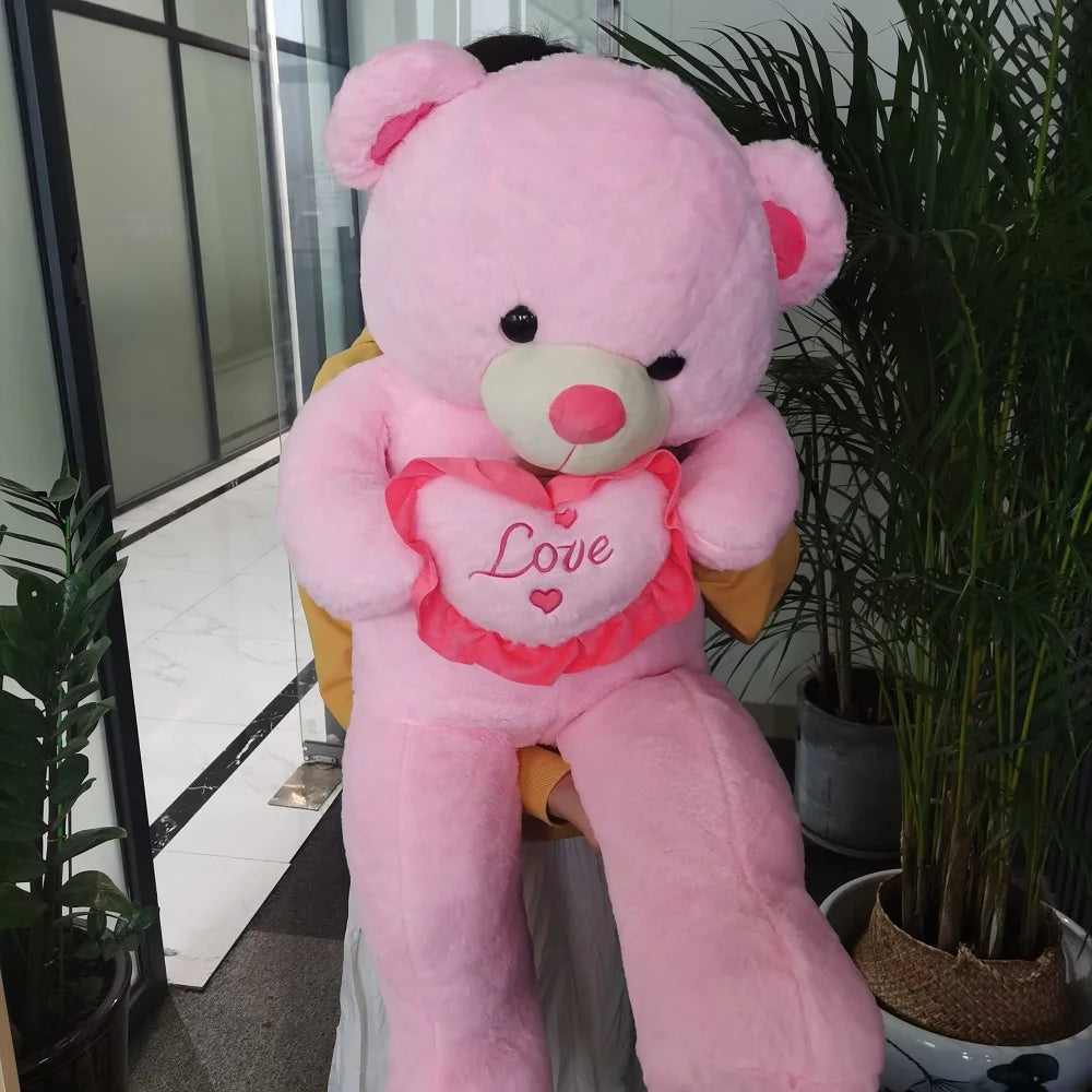 Large pink teddy bear with a heart that says 'Love' in an indoor setting with plants.