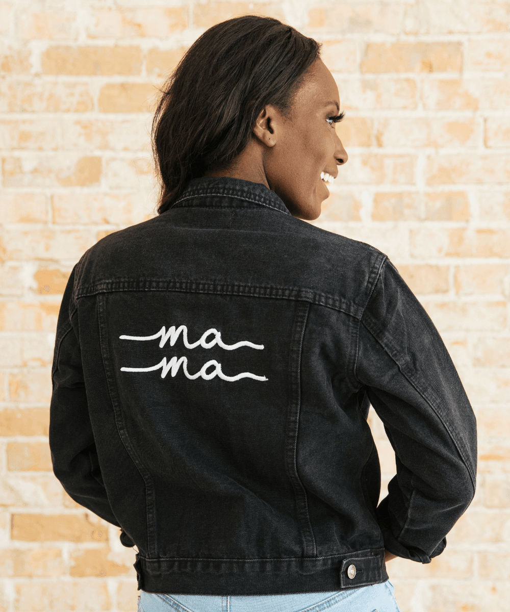 Person wearing a black denim jacket with 'ma ma' text on the back against a beige brick wall.