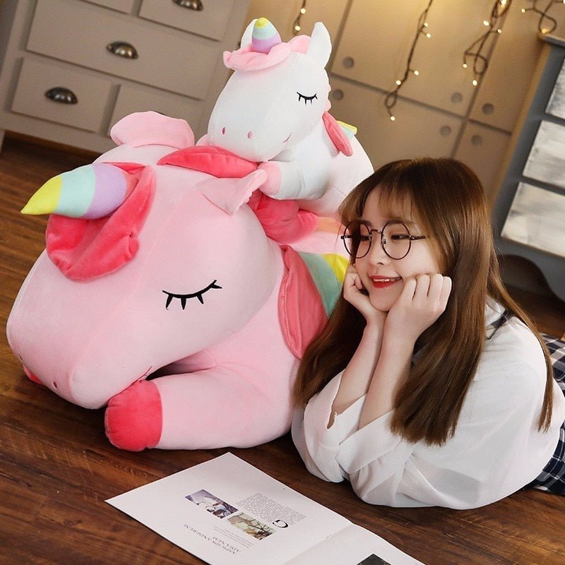 Person sitting next to a large pink and white unicorn plush toy on a wooden floor.