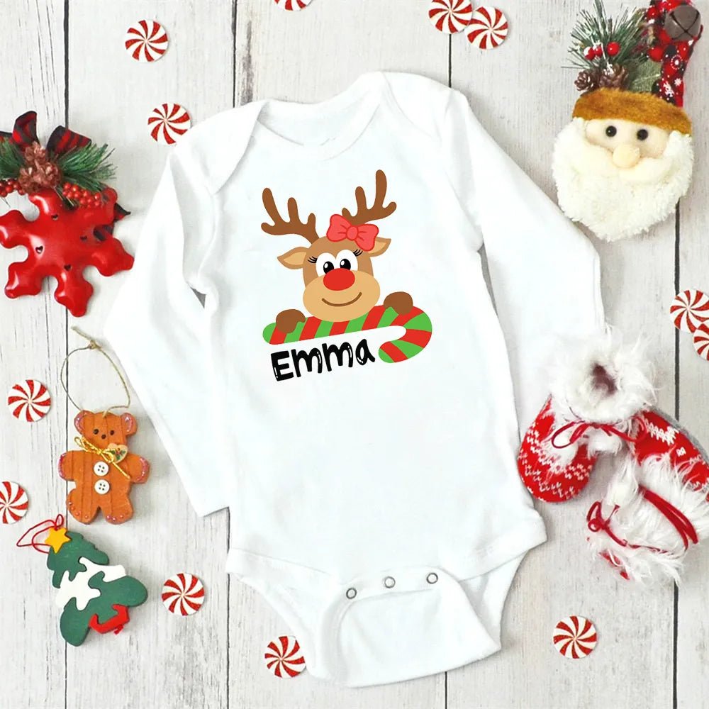 White baby onesie with a reindeer design and 'Emma' text, surrounded by Christmas decorations on a wooden surface.