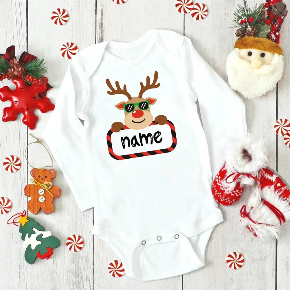 White baby onesie with reindeer design and 'name' text, surrounded by Christmas decorations on a wooden surface.