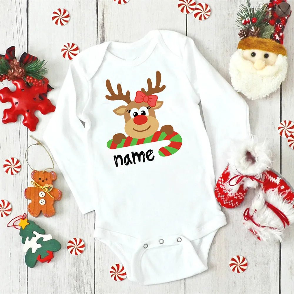 White baby onesie with a reindeer design and 'name' text, surrounded by Christmas decorations on a wooden surface.