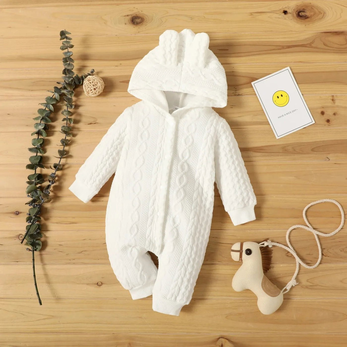 White baby snowsuit with cable knit pattern on a wooden surface with a toy and card.