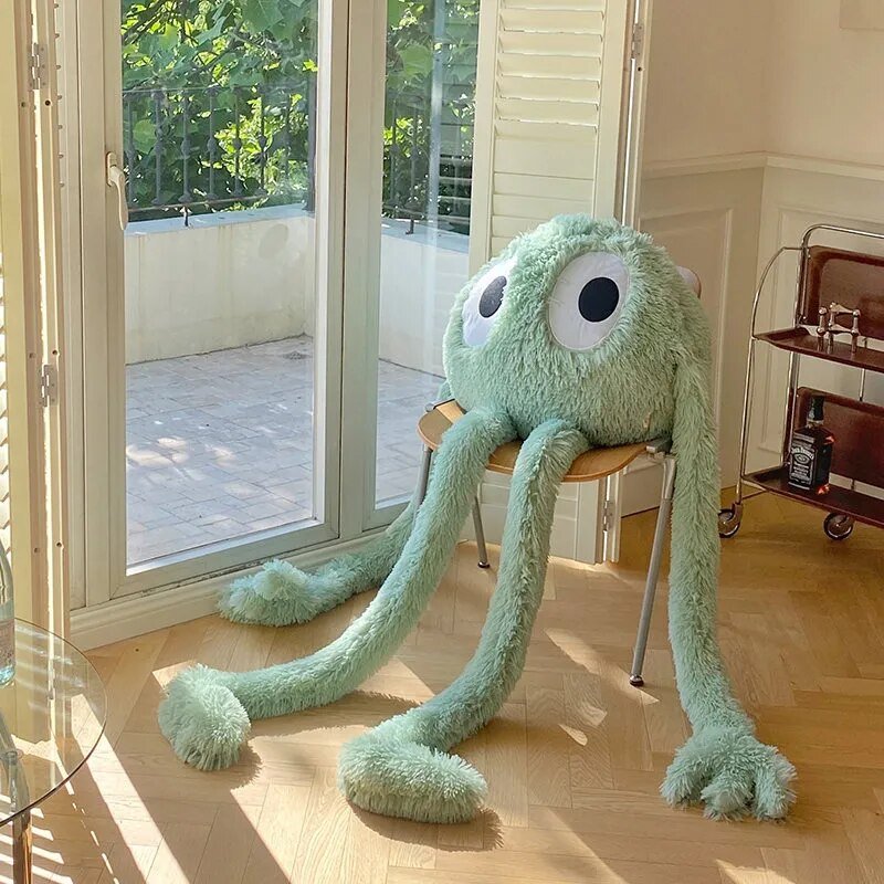Large green plush toy with long arms and legs on a chair in a room with large windows.