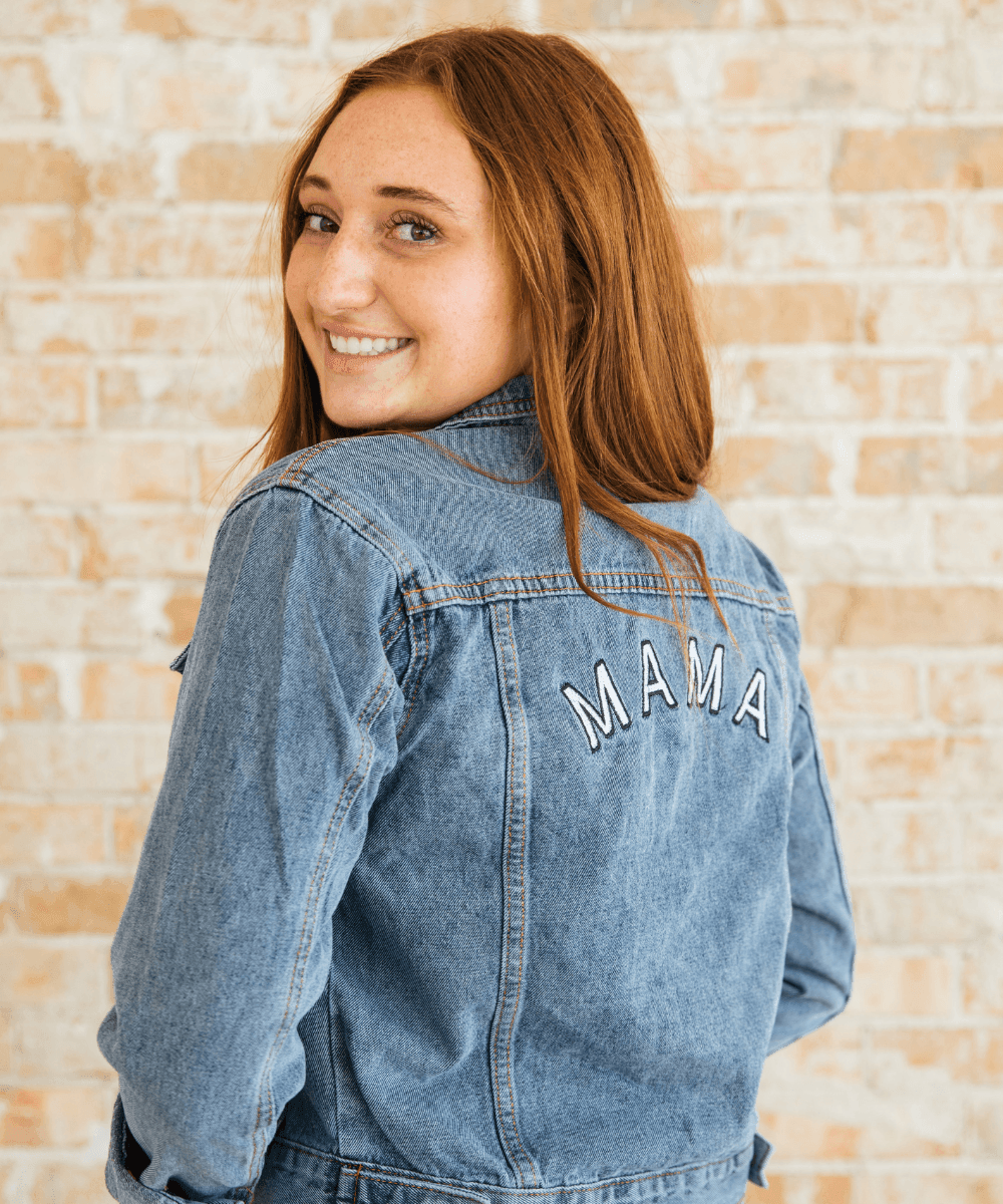 Person wearing a denim jacket with 'MAMA' on the back against a brick wall.