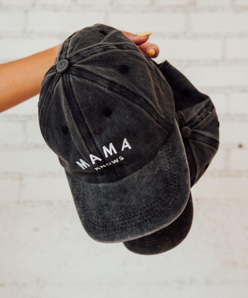 Black velvet cap with 'MAMA KNOWS' text held by a hand on a light wooden surface