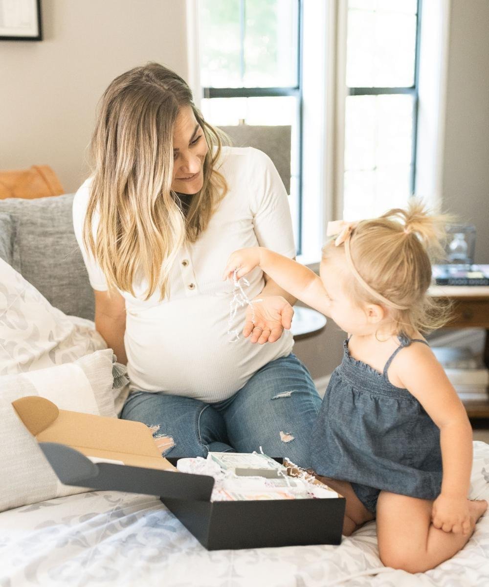 Pregnant woman sitting on a bed with a toddler, interacting with a box on the bed.