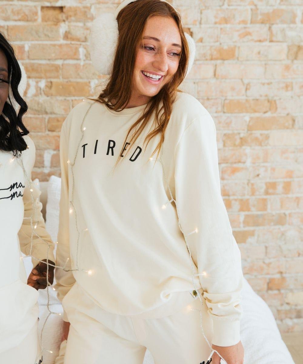 Woman wearing a cream-colored sweatshirt with 'TIRED' text, standing against a brick wall.