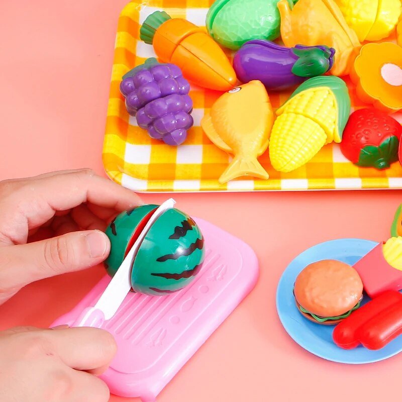 Colorful toy fruits and vegetables on a pink surface with a hand interacting with a toy watermelon.