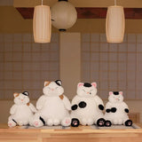 Four plush cat toys on a wooden surface with a tiled wall and hanging lights in the background.