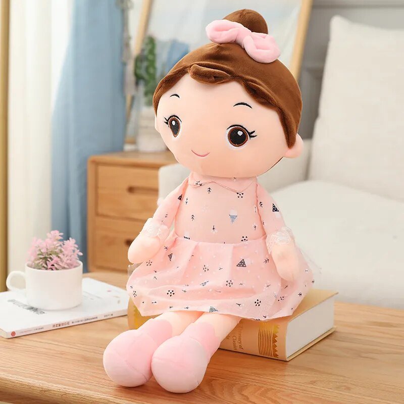 Plush toy of a girl in a pink dress with a bow on a wooden surface.