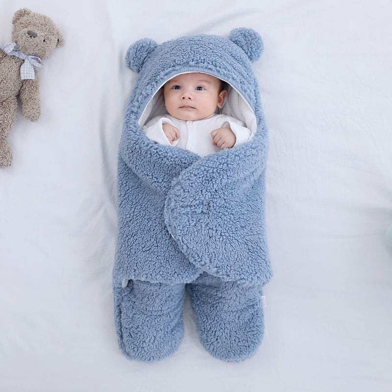 Baby wrapped in a blue fluffy blanket with bear ears, next to a teddy bear on a white background