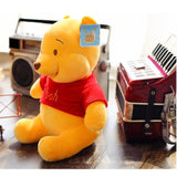 Plush toy bear wearing a red shirt with a tag, sitting on a wooden surface with musical instruments in the background.