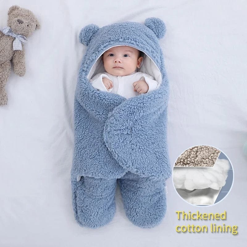 Baby wrapped in a blue fluffy swaddle with a teddy bear toy on a white background