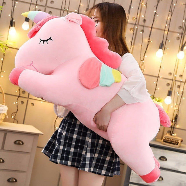 Person holding a large pink unicorn plush toy in a cozy room with string lights.