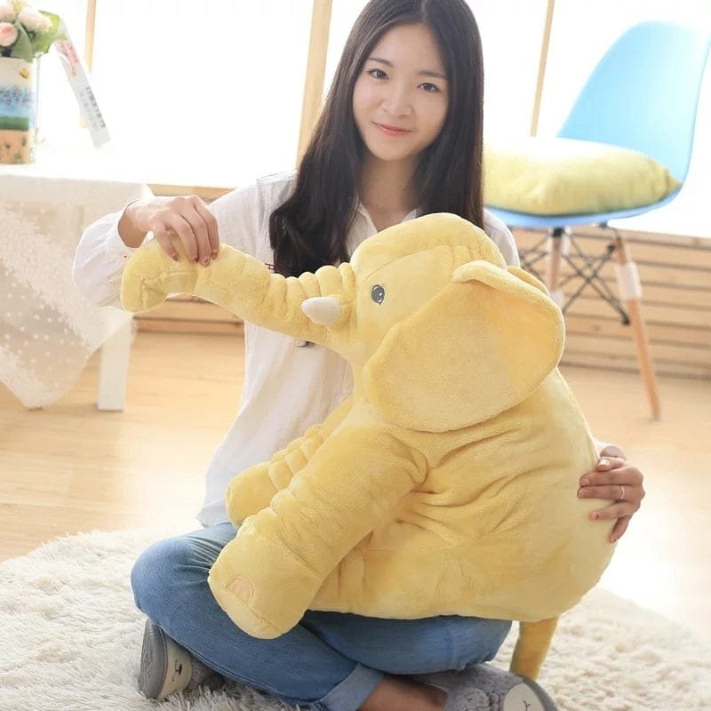 Woman holding a large yellow elephant plush toy in a room with a chair and plant.