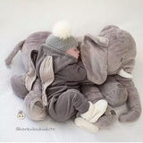 Child in a gray outfit lying next to a large plush elephant toy on a white background