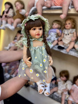 Doll in a green dress with floral patterns held by a person, with other dolls in the background.