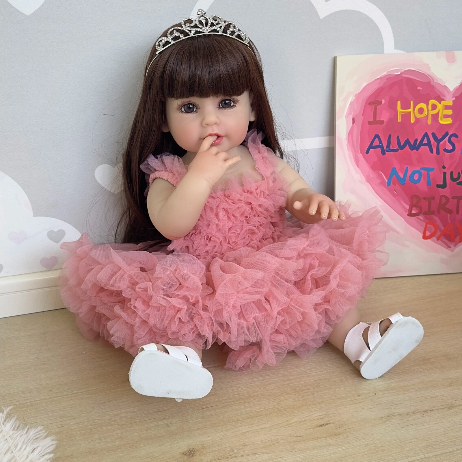 Realistic full body silicone Reborn Toddler Doll in a pink princess dress seated on wooden flooring for nursery display and pretend play.