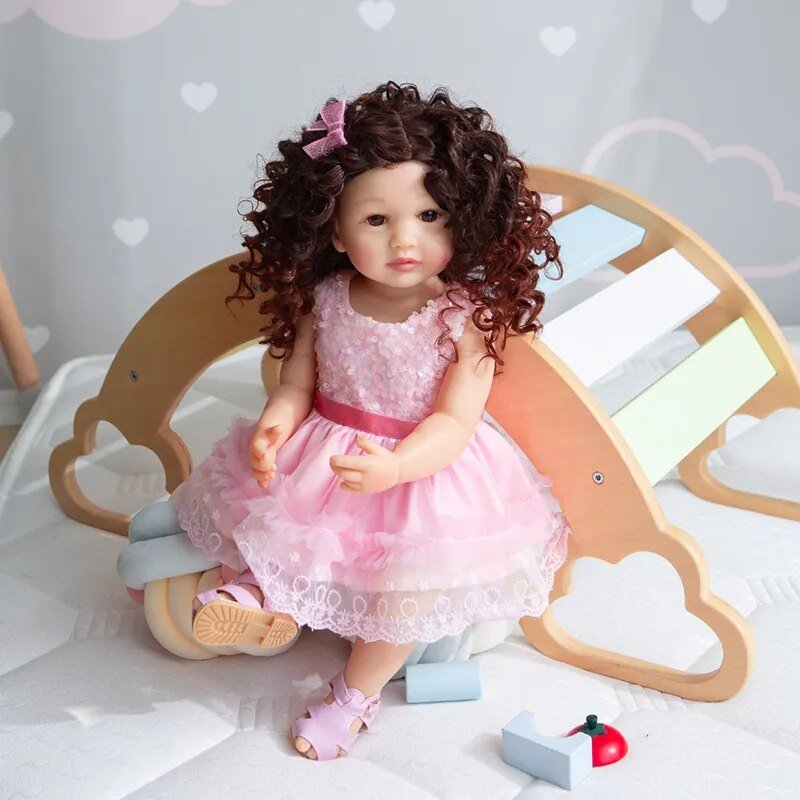 Soft touch silicone Reborn Toddler fashion doll in a pink dress seated on wooden flooring for nursery display and pretend play.