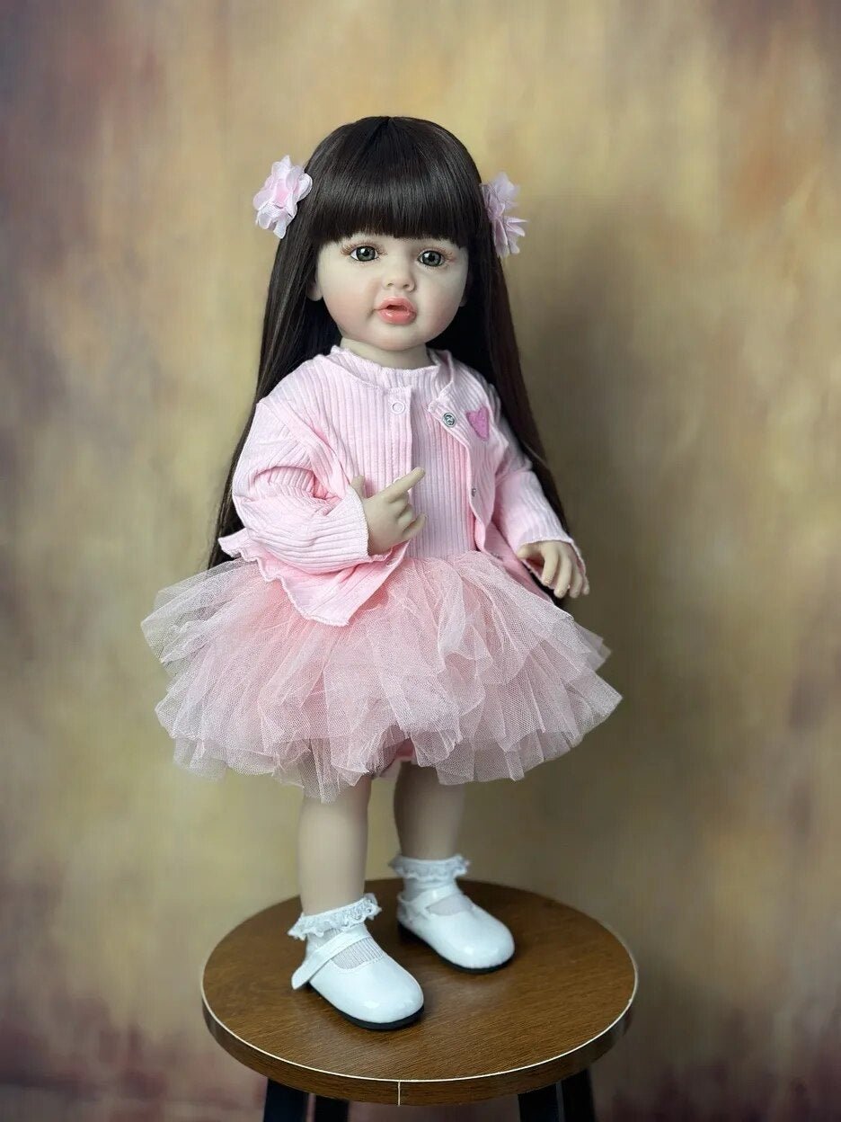 Standing Reborn Doll in a pink tutu skirt seated on a wooden stool, a lifelike silicone doll for dress-up play and shelf display.
