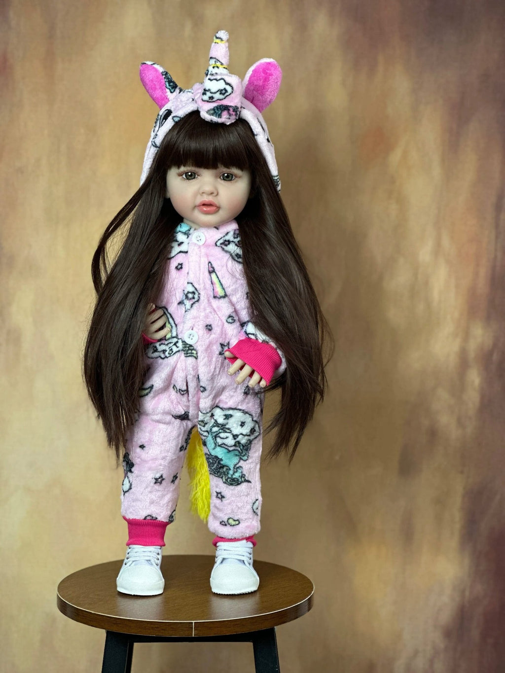 Close-up of the Standing Reborn Doll in a unicorn onesie on a wooden stool, a realistic silicone doll for cosy pretend play.