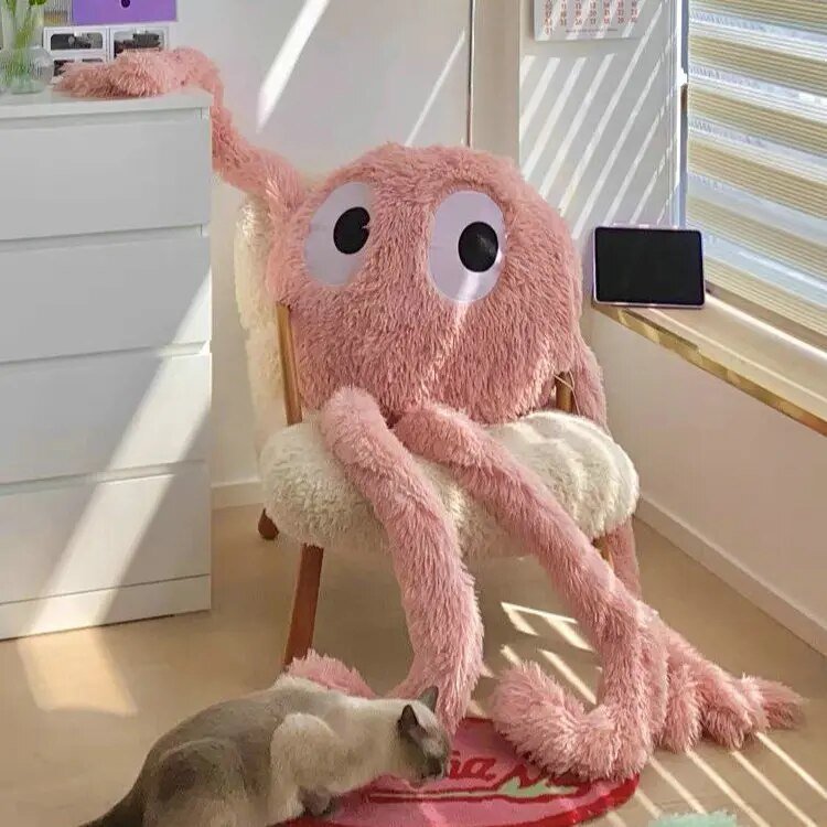 Pink plush toy with googly eyes on a chair, with a cat interacting with it in a room.