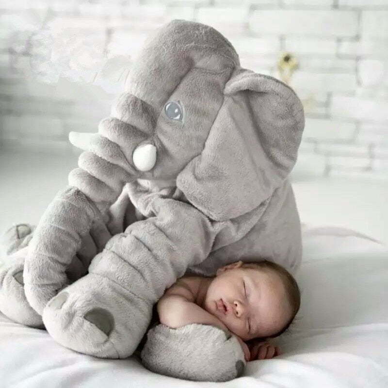 Baby sleeping under a large gray elephant plush toy on a white blanket.