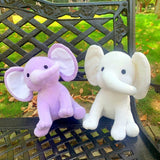 Two elephant plush toys, one purple and one white, sitting on a black metal chair outdoors.