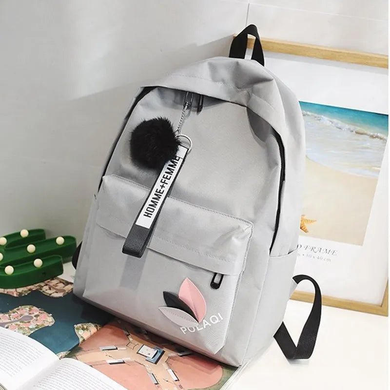 Gray backpack with a pom-pom keychain on a surface with a magazine and green object in the background.