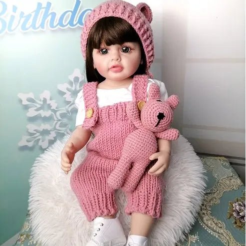A child-like doll wearing a pink knitted outfit with a bear toy.
