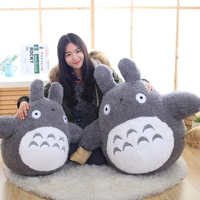 Person sitting with two large plush Totoro toys on a wooden floor.