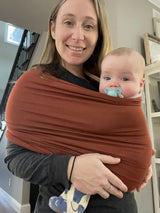 Woman holding a baby wrapped in a brown sling at home.