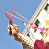 Person holding a pink and orange slingshot against a clear blue sky.