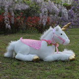 Plush unicorn toy with pink saddle blanket on grass with flowers.