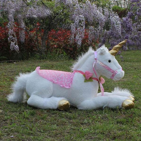 Plush unicorn toy with pink saddle blanket on grass with flowers.