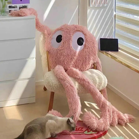 Pink plush toy with googly eyes on a chair, with a cat interacting with it in a room.