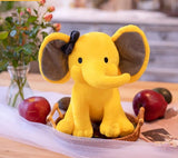 Yellow elephant plush toy with a black bow on a table with fruits and flowers.