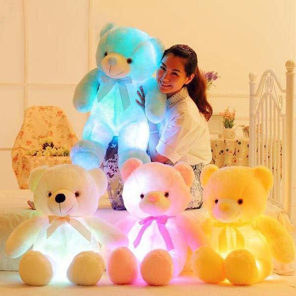 Woman holding a glowing teddy bear with three other colorful teddy bears on a bed.
